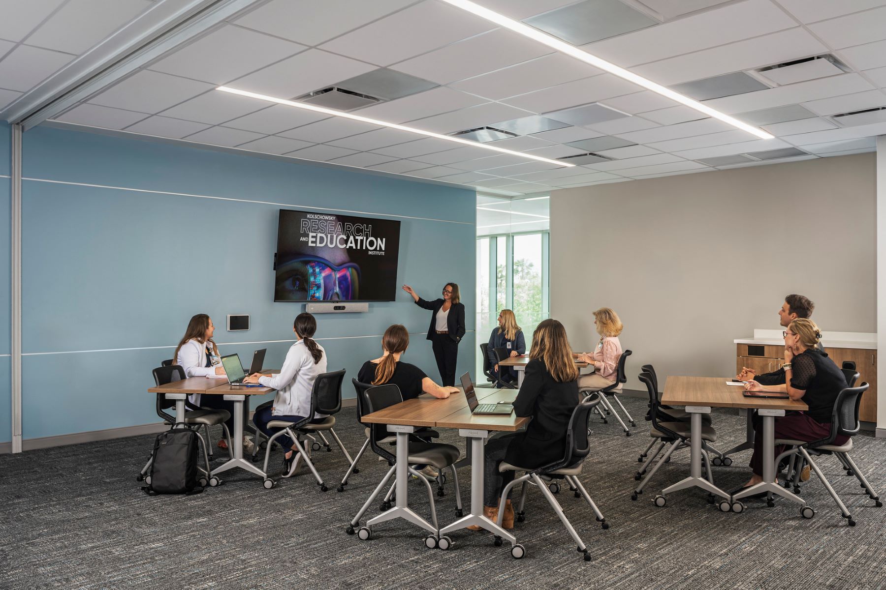 person standing and pointing at a monitor with six people seated at desks