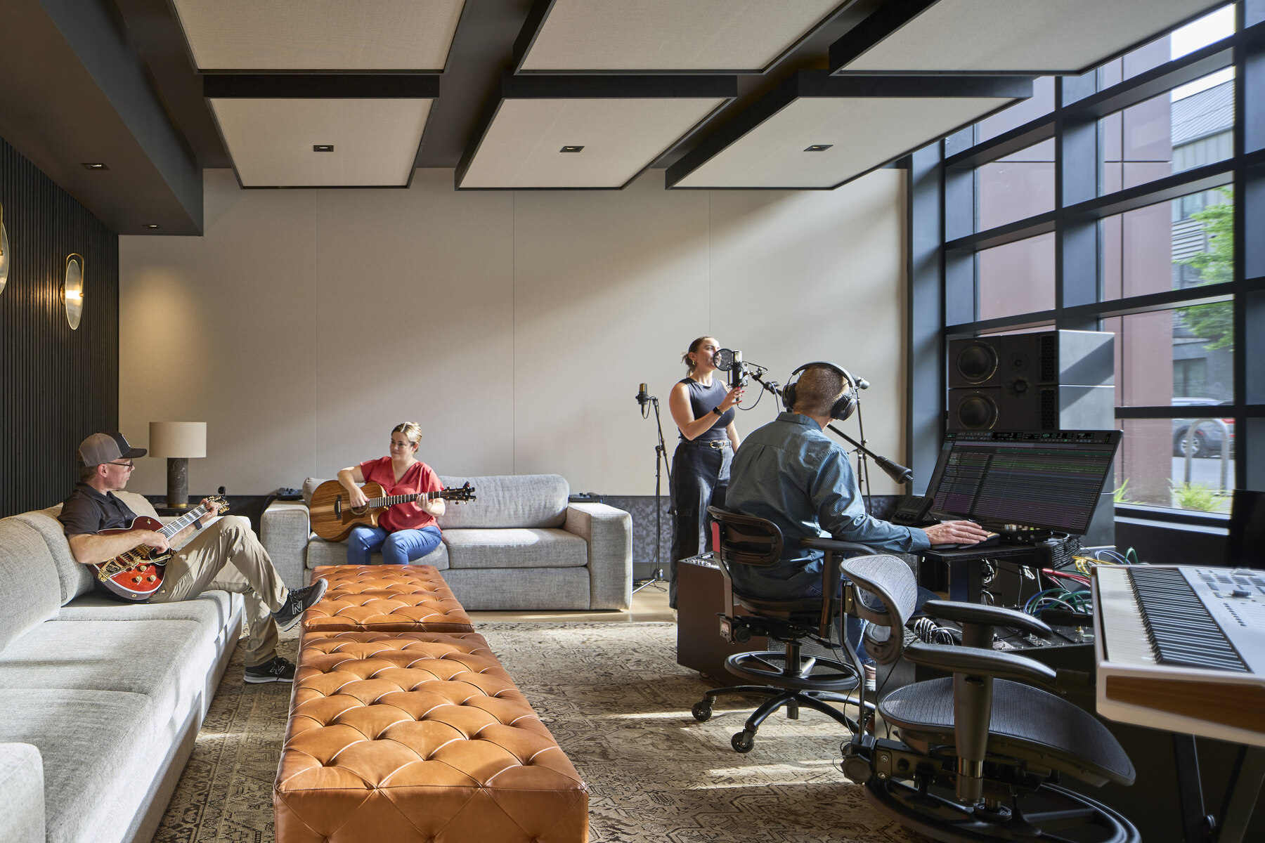 Two people on a couch playing guitar, one person at a computer and one person standing at a microphone in a music studio